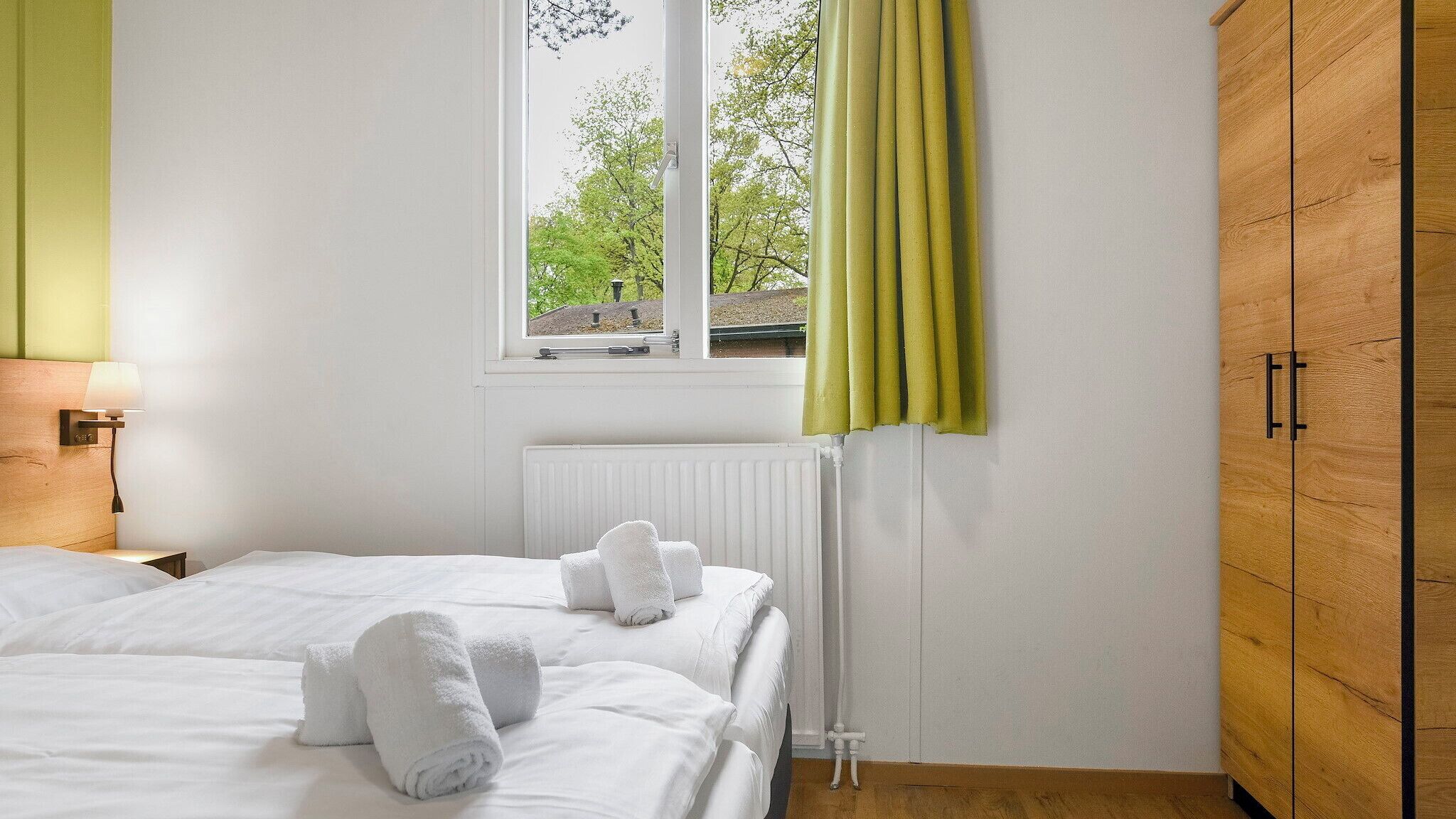 Ein Hotelzimmer mit zwei Betten, einem Fenster mit Vorhängen, einem Heizkörper und einem Holzschrank.