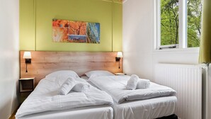 A double room with two beds, a wooden headboard, bedside lamps, a window with a view of trees, and a colorful artwork on the wall.