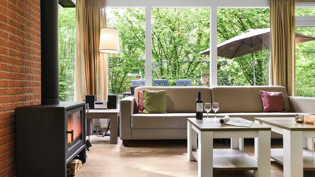 A cozy living room with a fireplace, a sofa, a coffee table, and a large window with curtains. - Comfortable 8-person bungalow (Putten)