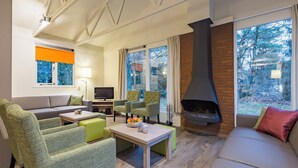 Una sala de estar moderna con chimenea, muebles verdes y grises, televisor y ventanas amplias con vistas a árboles.