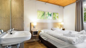 A hotel room with a wooden ceiling, a white sink, a bed with white linens, two bedside lamps, and three wall-mounted paintings.