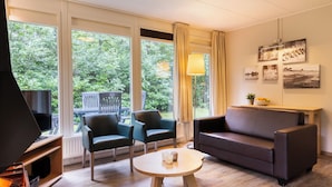 A modern living room with a sofa, two armchairs, a coffee table, and a TV. Large windows offer a view of greenery outside. - Comfortable bungalow for 4 persons (Putten)