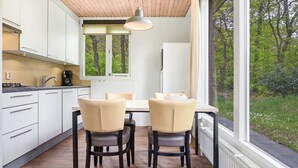 A modern kitchen with white cabinets, a dining table with two chairs, and a large window offering a view of trees. - Semi-detached bungalow for 4 persons (Putten)