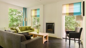 A living room with a green sofa, a grey sofa, a wooden coffee table, a fireplace, and large windows with curtains. - Markegaerde Wellness 4 (Ruinen)