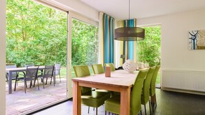 A dining area with a wooden table and green chairs, a hanging lamp, and a view of a lush garden through large windows. - Markegaerde Wellness 4 (Ruinen)
