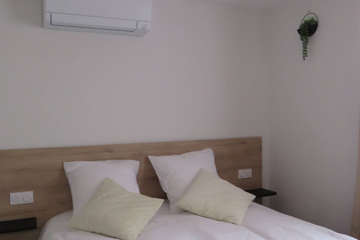 A hotel room with a bed, white linens, two pillows, a headboard, an air conditioning unit, a bedside table, and a wall-mounted plant.