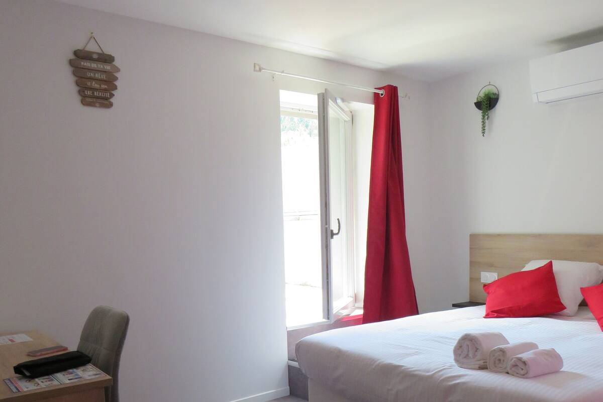 A hotel room with a bed, a desk, a chair, a red curtain, and a small plant on the wall.