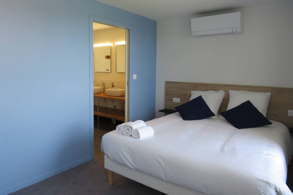 A hotel room with a bed, a nightstand, a wall-mounted air conditioner, and a bathroom visible through an open door.