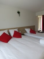 Habitación de hotel con dos camas, cada una con ropa blanca y almohadas rojas. Hay una planta pequeña colgada en la pared, encima de las camas.