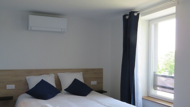 A neatly made bed with pillows, a bedside table, and a window with curtains. - Les Jardins de Saulzet (Romagnat)