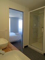 Un baño moderno con una bañera blanca grande, cabina de ducha con paredes de vidrio y una cama visible a través de una puerta abierta.