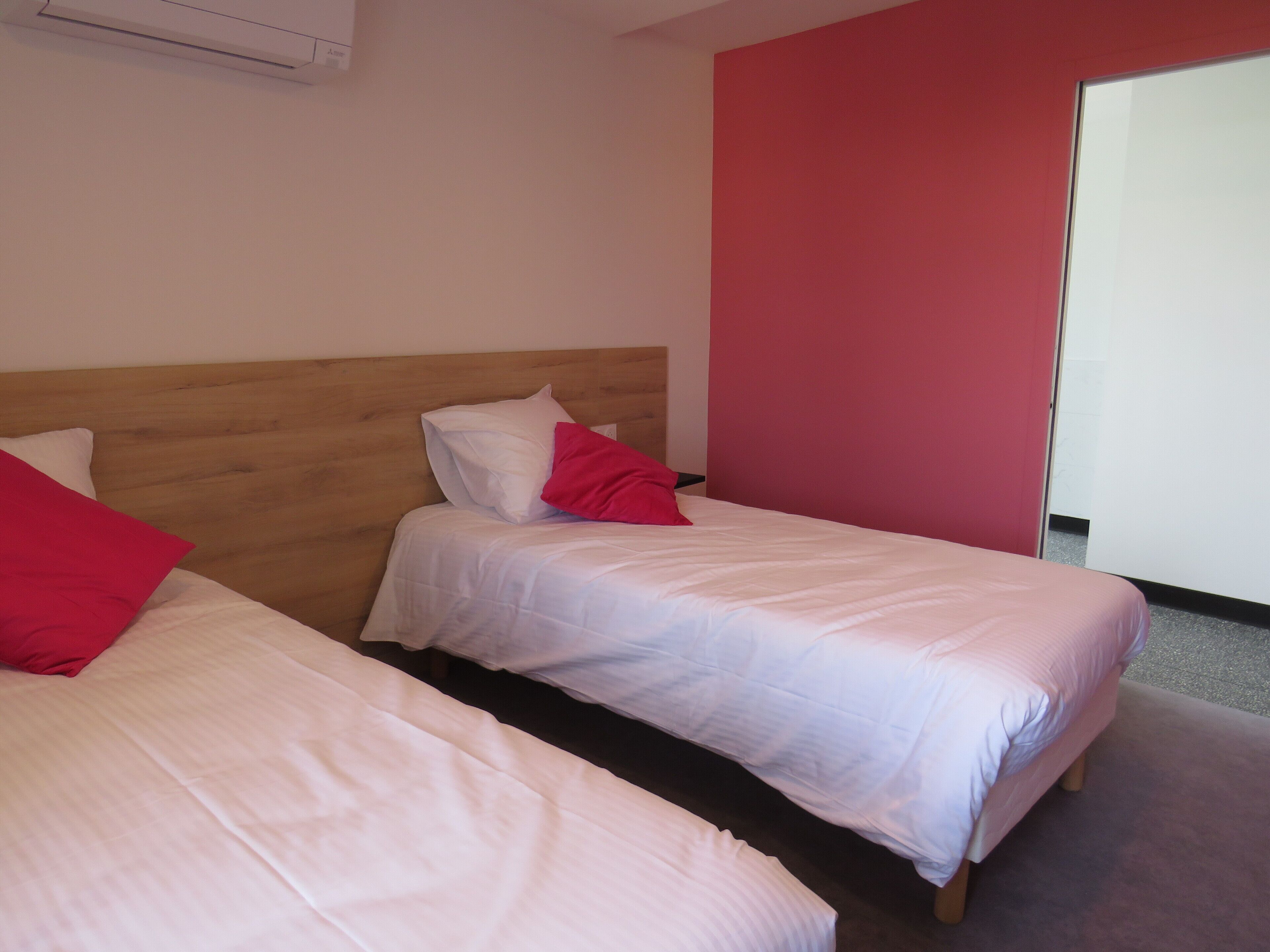 Dos camas individuales con cabeceros de madera, ropa de cama blanca y almohadas rojas en una habitación con una pared de acento roja y una puerta que da a otra habitación.