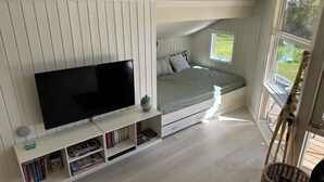 Une pièce comprenant un lit, un téléviseur posé sur un meuble, une bibliothèque et une fenêtre donnant sur un paysage verdoyant.