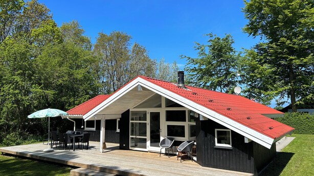 Une maison moderne avec un toit rouge, de grandes fenêtres et une terrasse aménagée avec du mobilier d’extérieur.