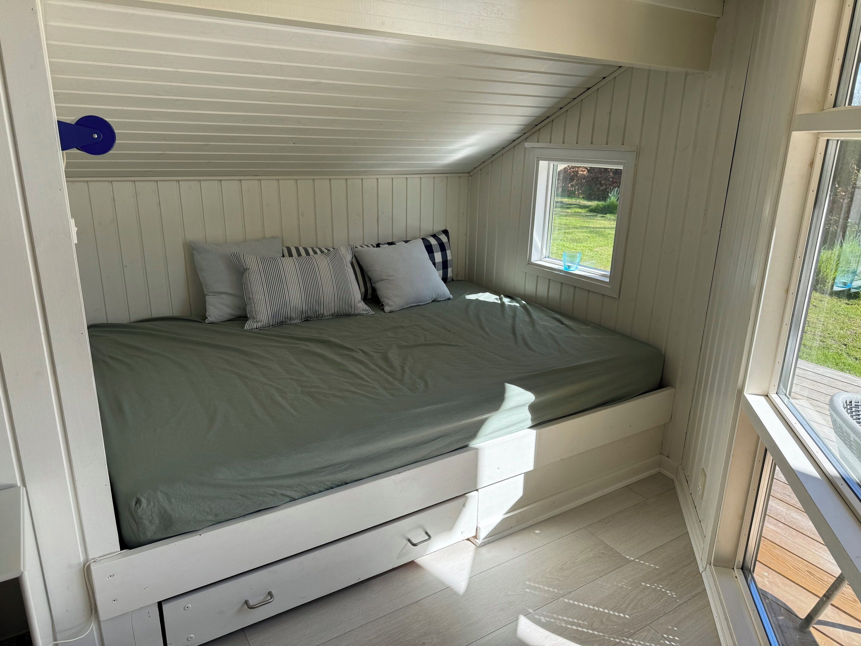 A single bed with a green mattress, white headboard, and a small window with a view of greenery.
