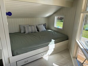 Un lit simple avec un matelas vert, une tête de lit blanche et une petite fenêtre donnant sur un paysage verdoyant.