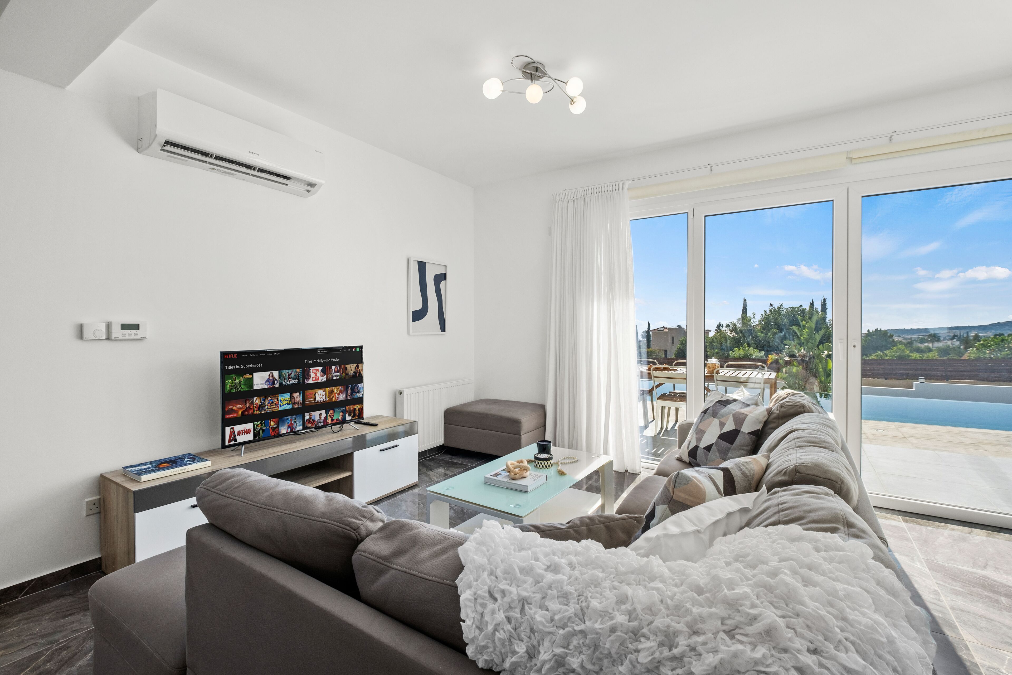 A modern living room with a sofa, a coffee table, and a TV. There is a large window with a view of a pool and greenery.