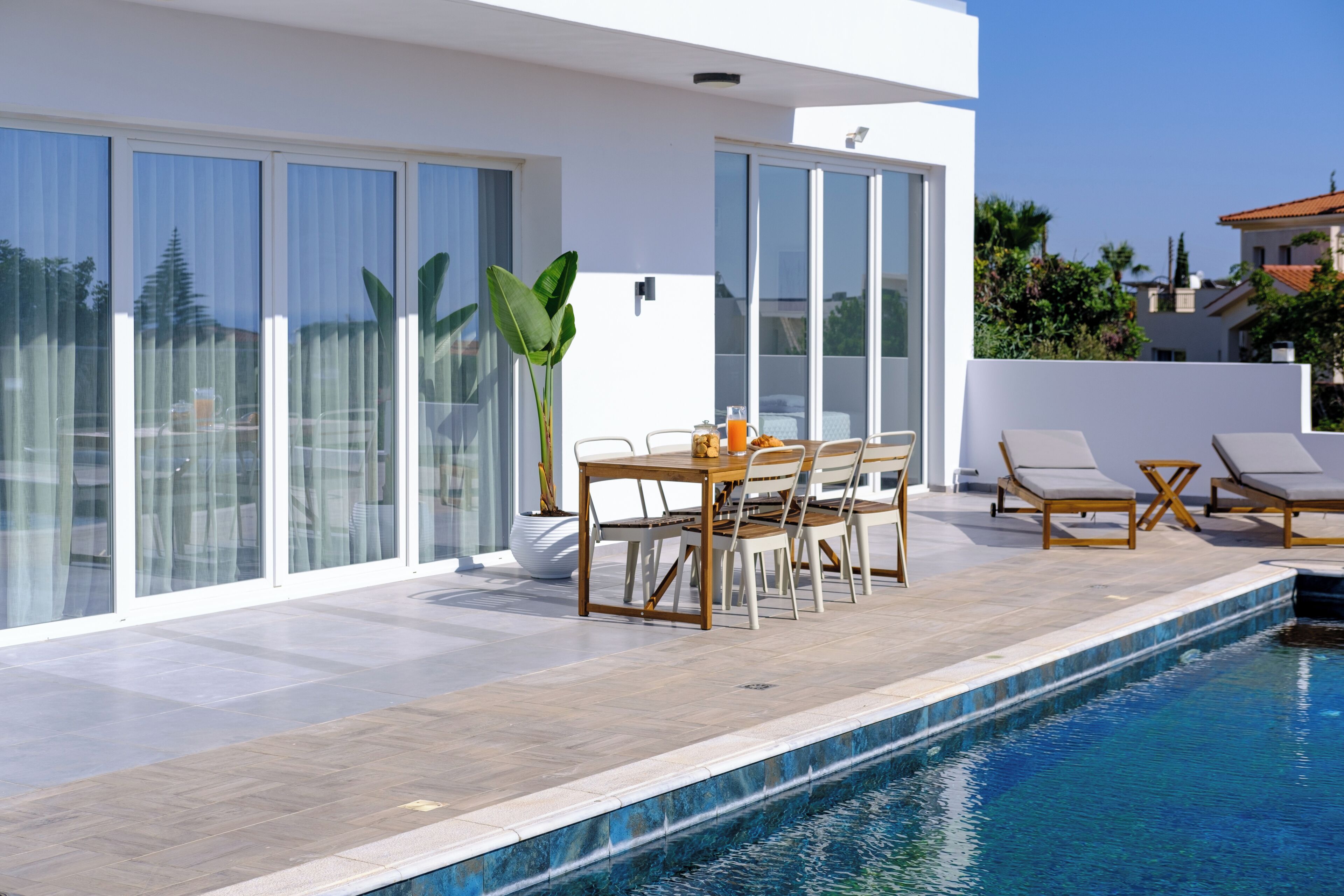 A modern outdoor pool area with a dining table and chairs, surrounded by large sliding glass doors and a view of the neighborhood.