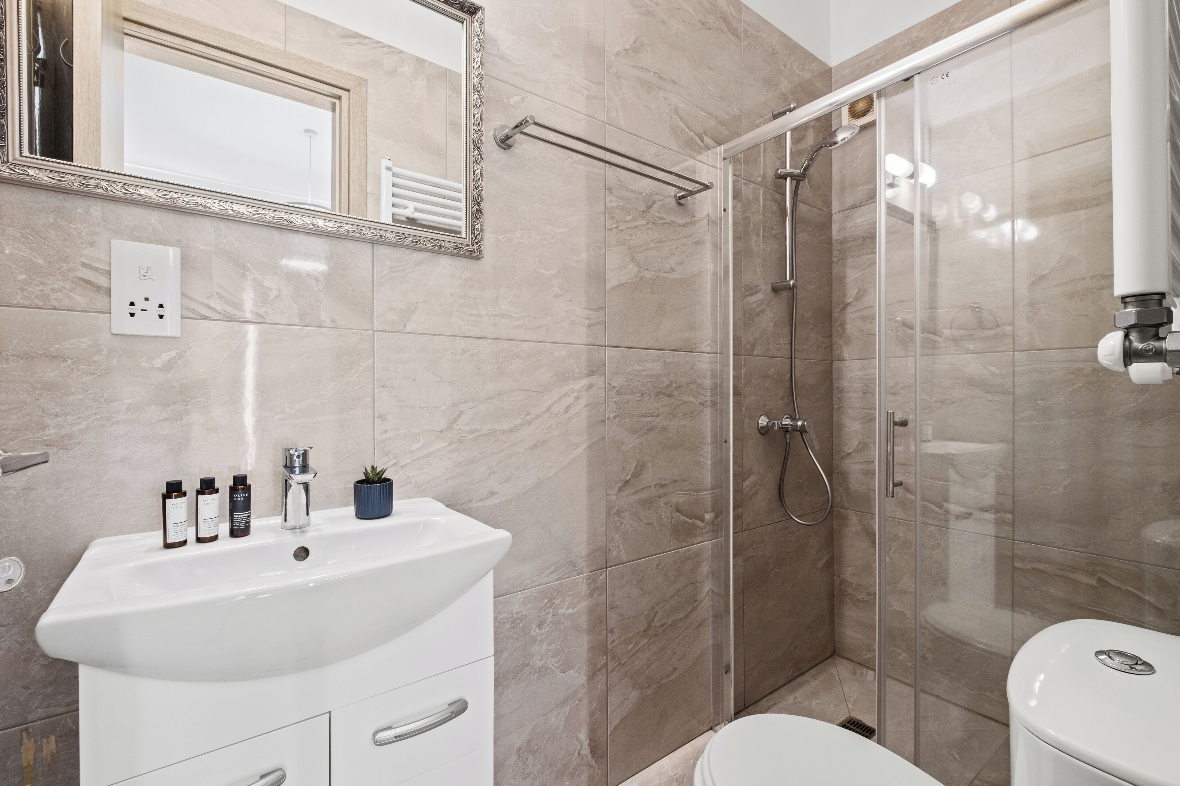 A modern bathroom with a white sink, mirror, and toilet. It includes a glass shower enclosure and a marble-tiled wall.