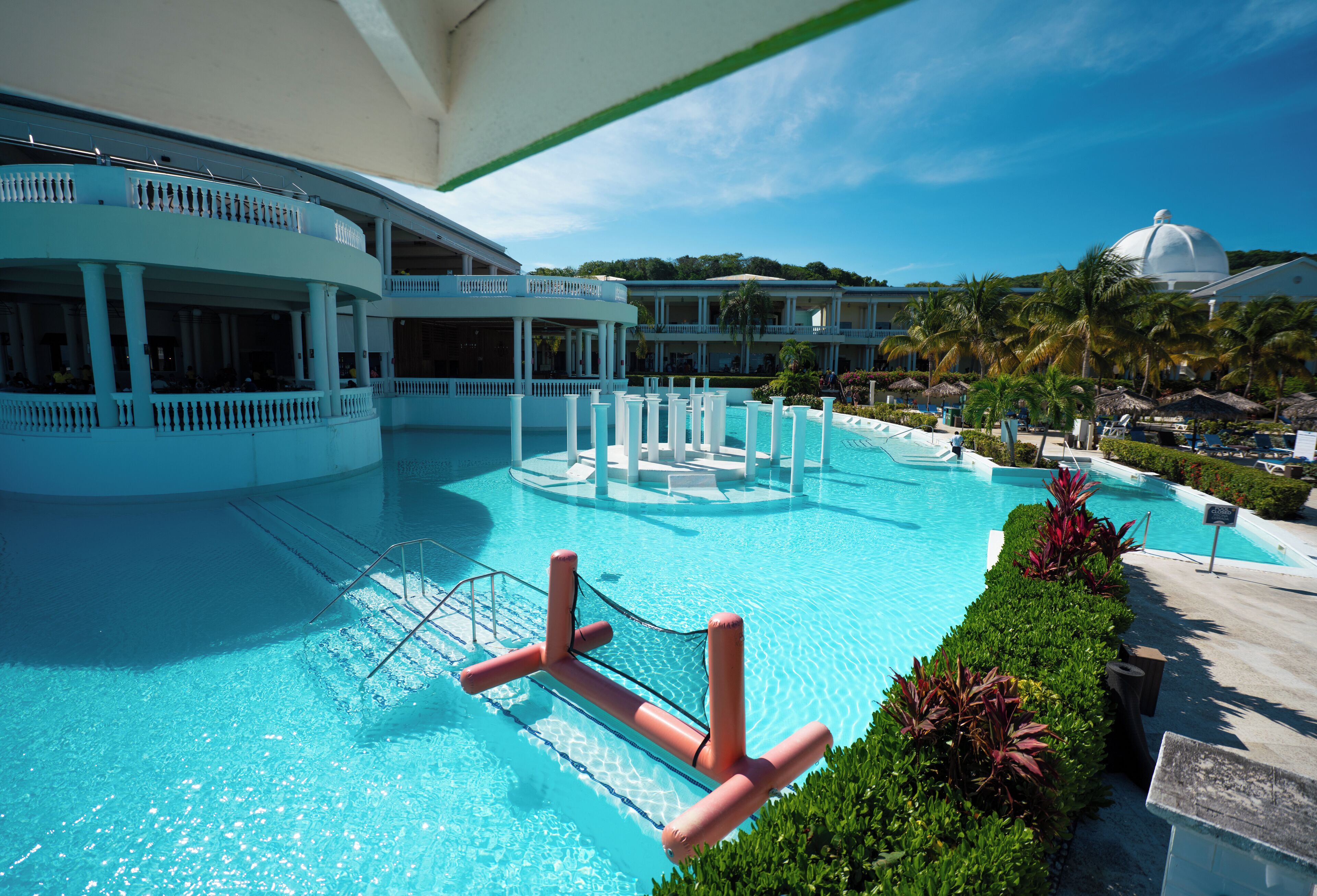A large swimming pool with a diving board, surrounded by lush greenery and a multi-story building with balconies.