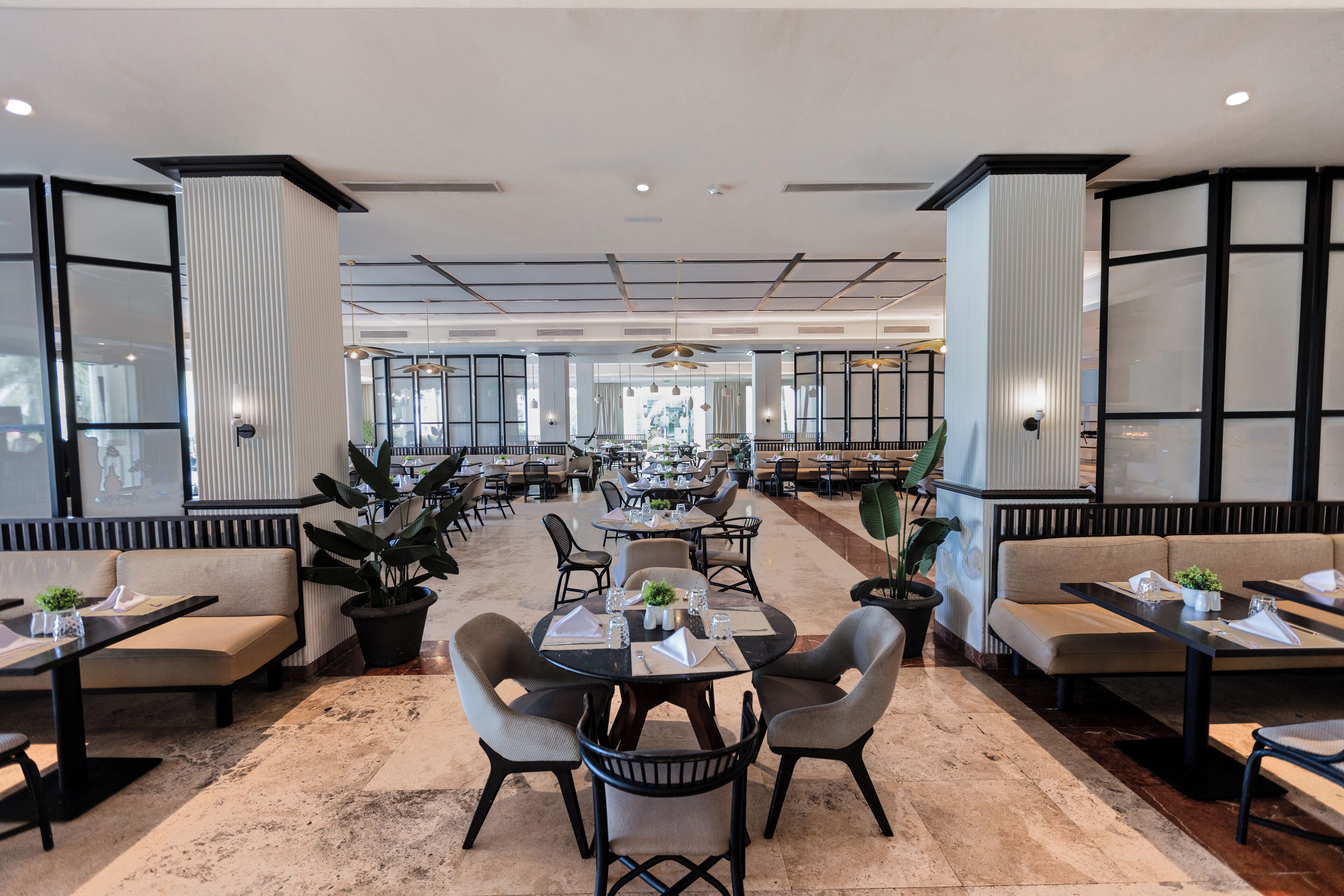 A spacious hotel lobby with multiple seating areas, including round tables and sofas, and large windows providing natural light.