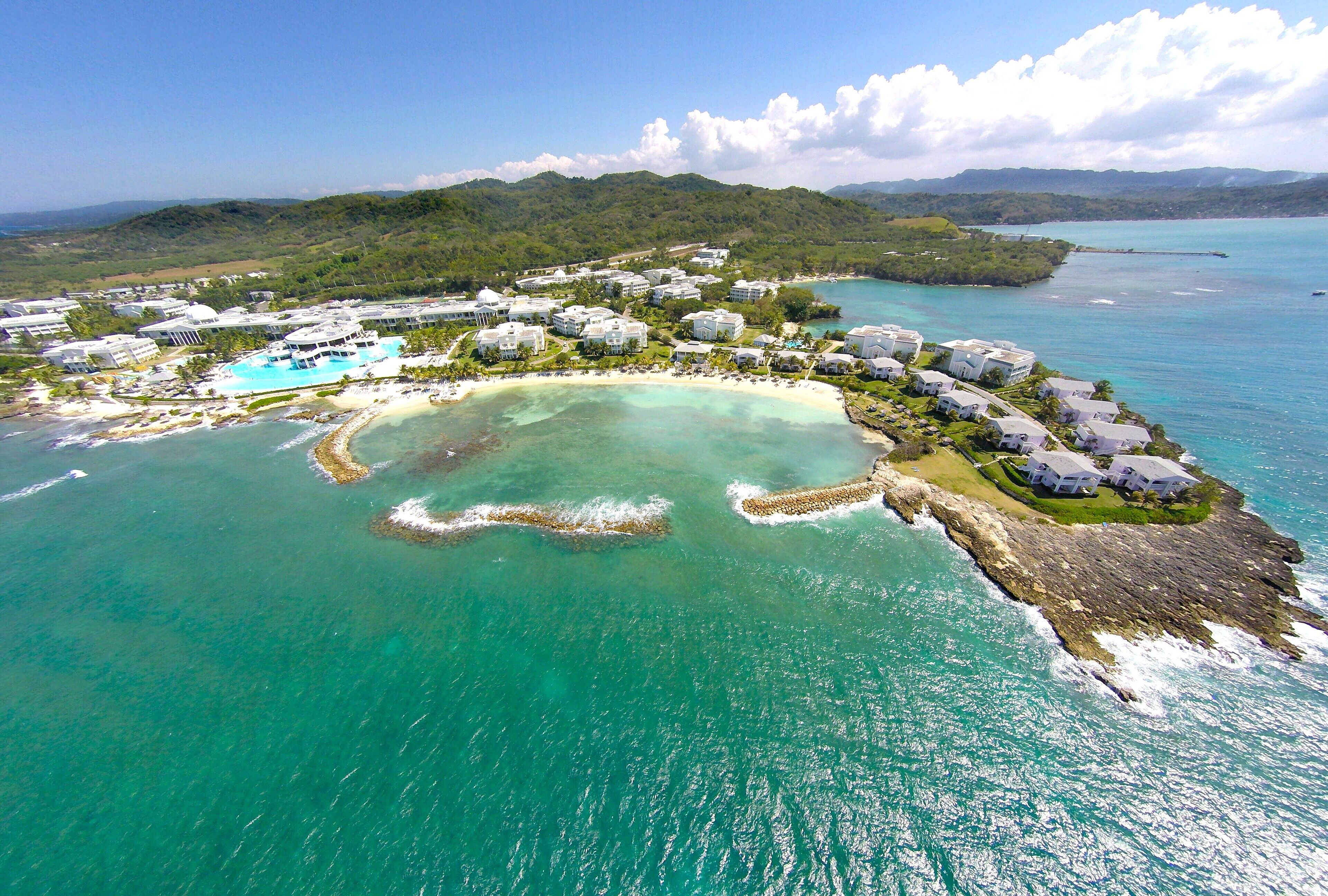 An aerial view of a coastal resort with multiple buildings, a central pool, and a beach.
