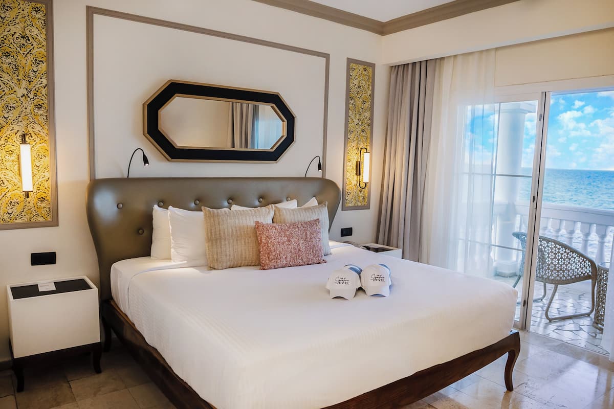A hotel room with a large bed, a nightstand, a mirror, and a view of the sea through a balcony door.