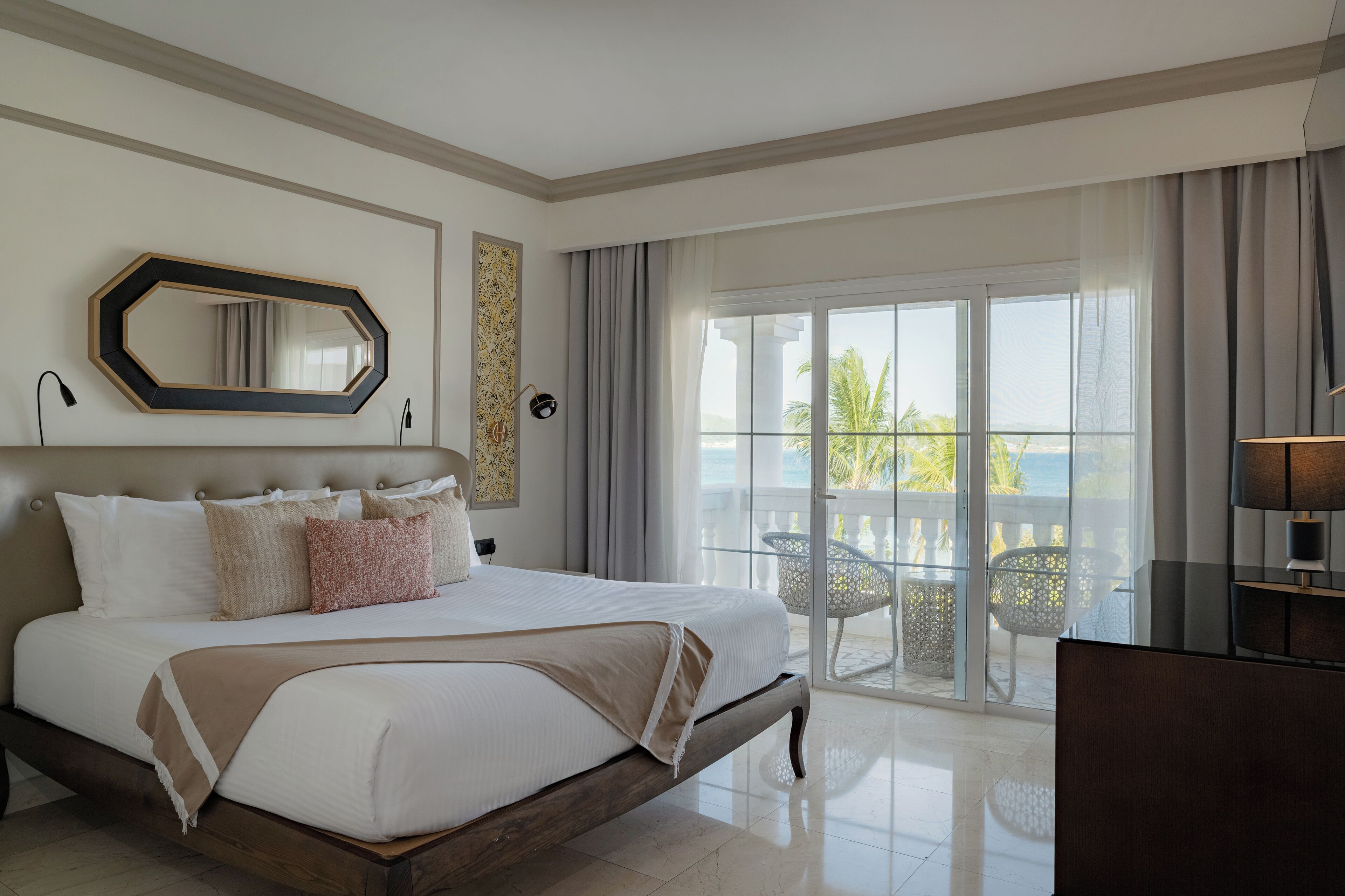 Une chambre d’hôtel avec un grand lit, un balcon avec des chaises, une vue sur la mer et un grand miroir au mur.