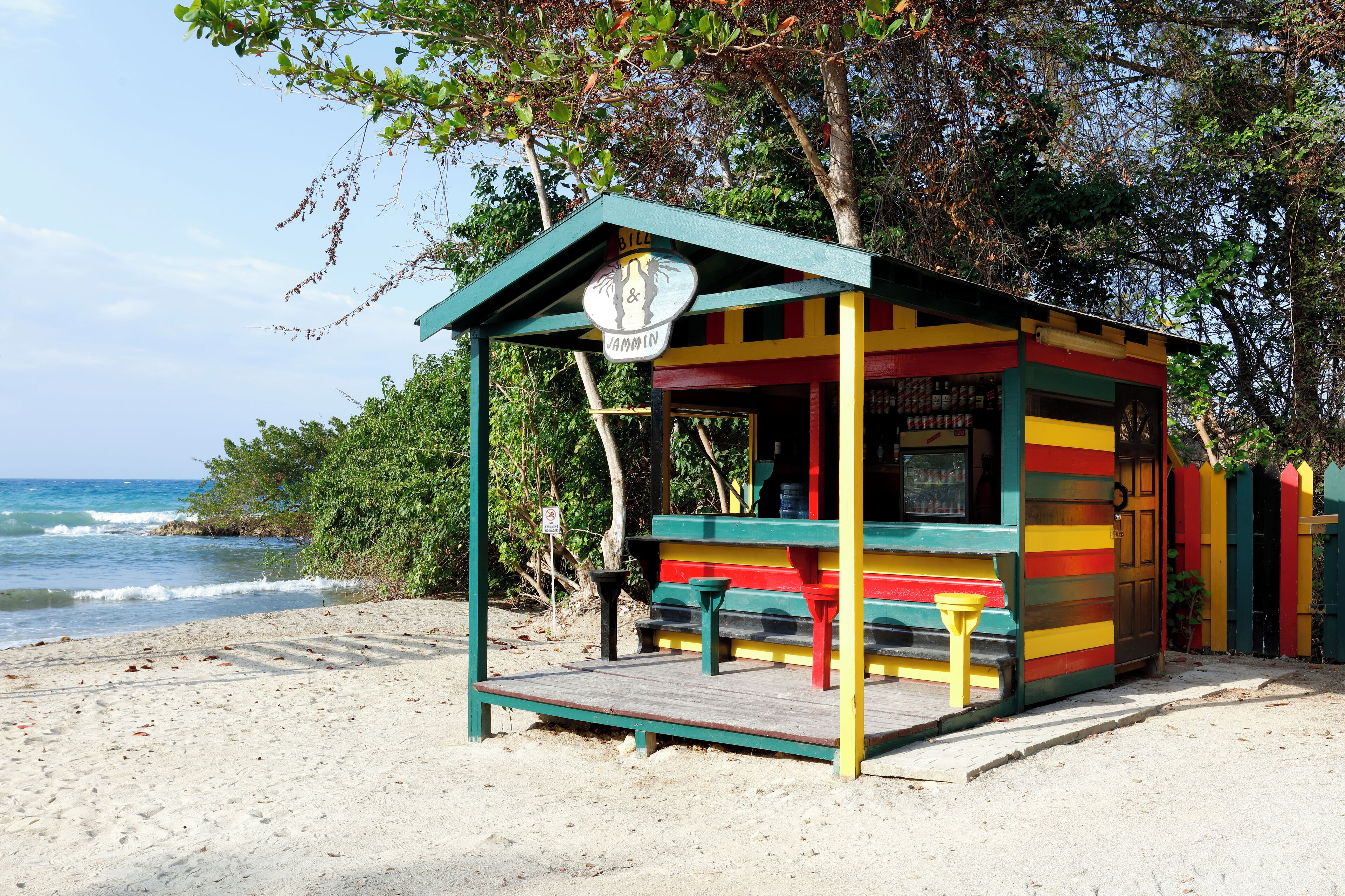 Un petit kiosque situé au bord de la plage, doté d’un toit végétalisé et de murs aux couleurs vives, avec une enseigne et une petite table.