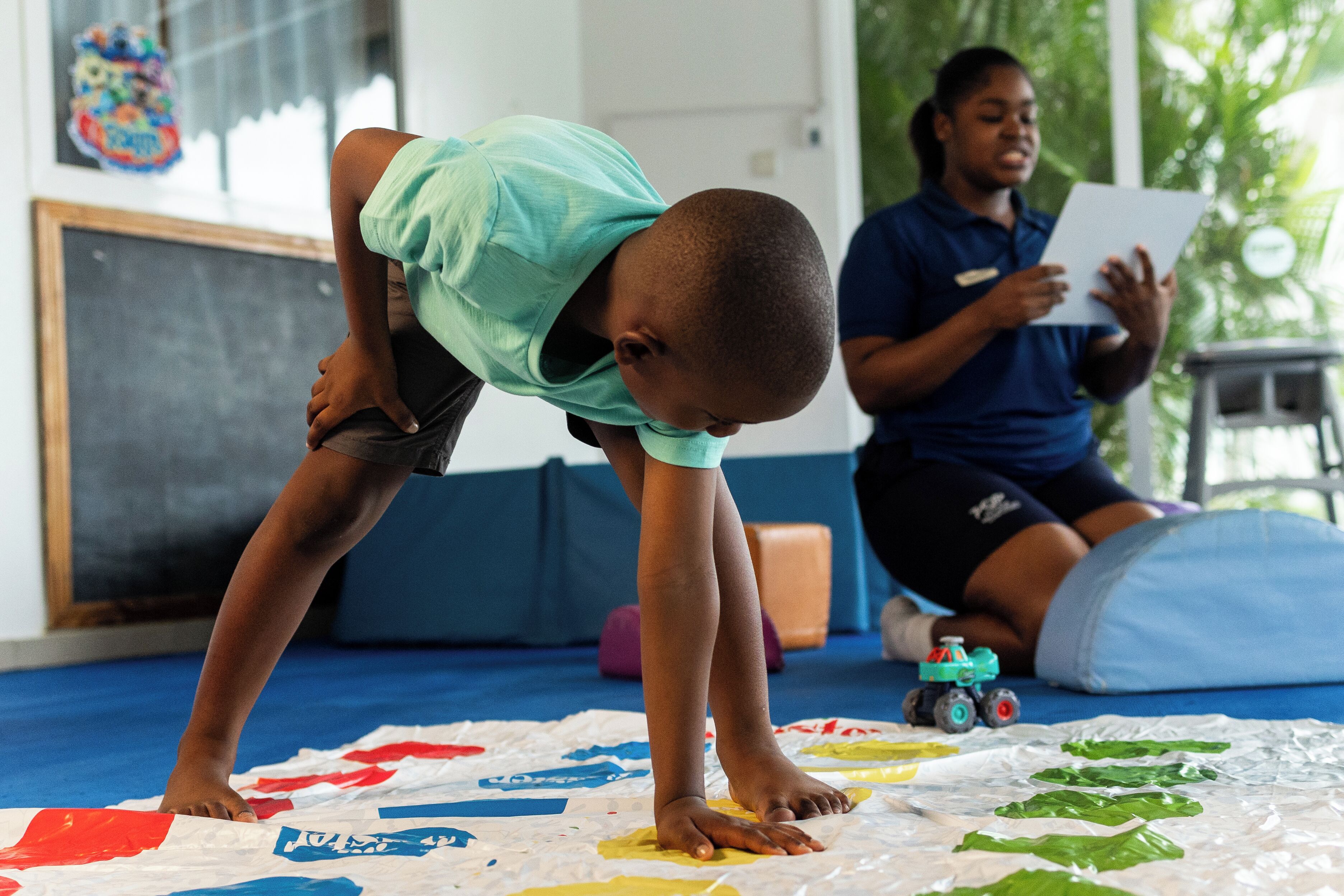 A child stretching on a mat with colorful paper on the floor, a small toy car, and a person sitting in the background.