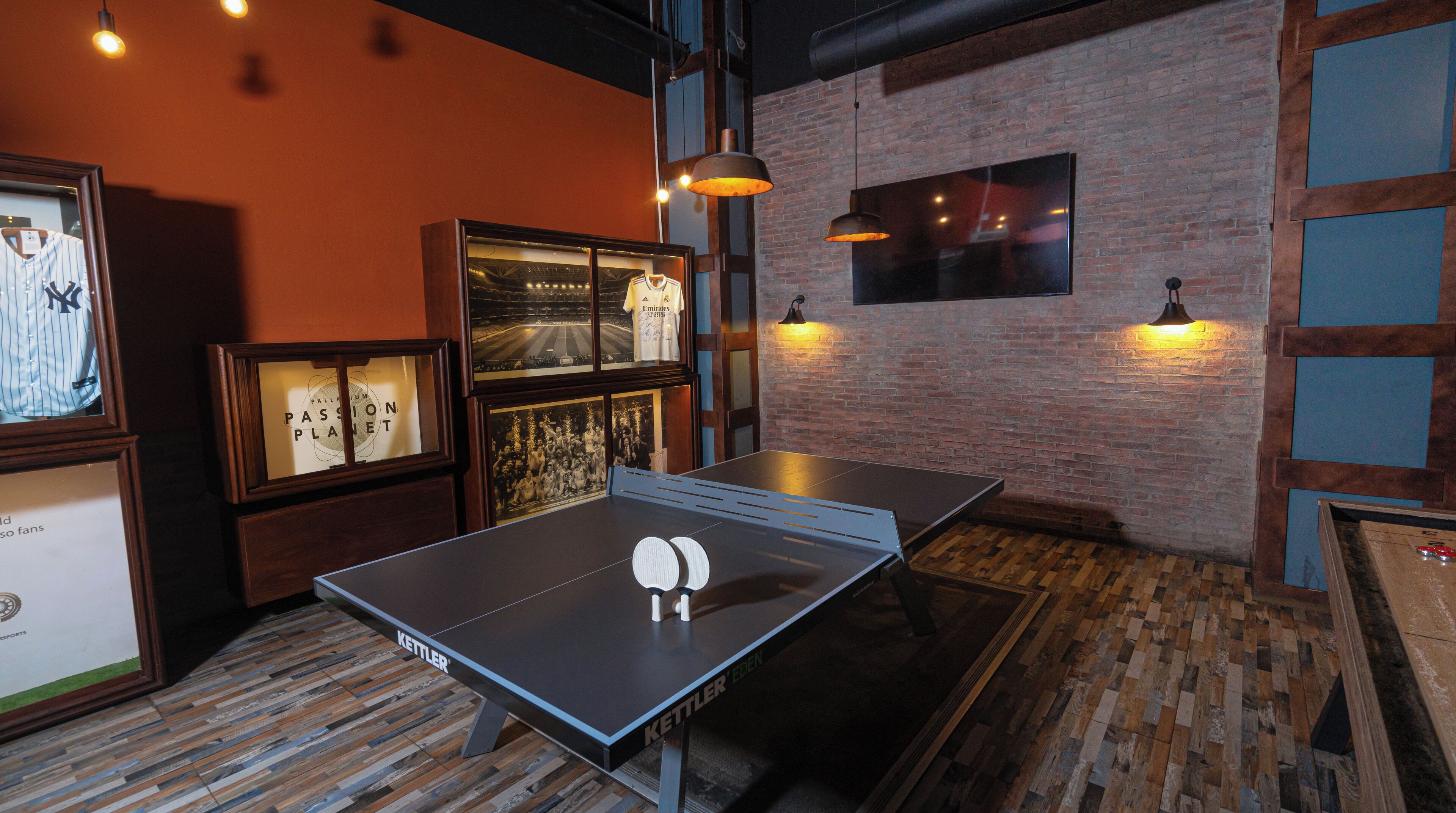 Una mesa de ping pong en una habitación con pared de ladrillo, cuadros enmarcados e iluminación colgante.