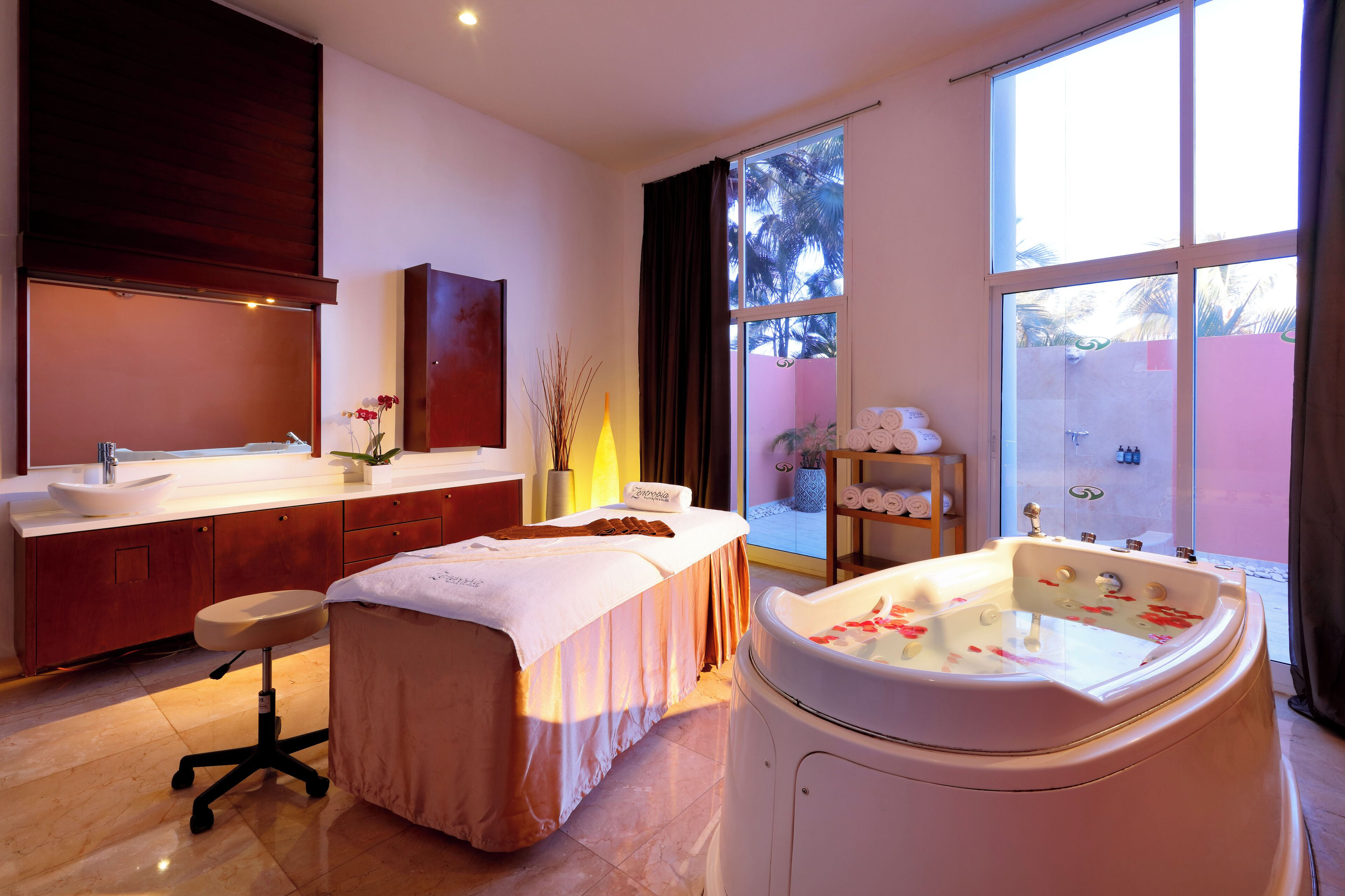 Un moderno spa con una bañera grande, una camilla de masaje y un amplio ventanal con vistas a palmeras.