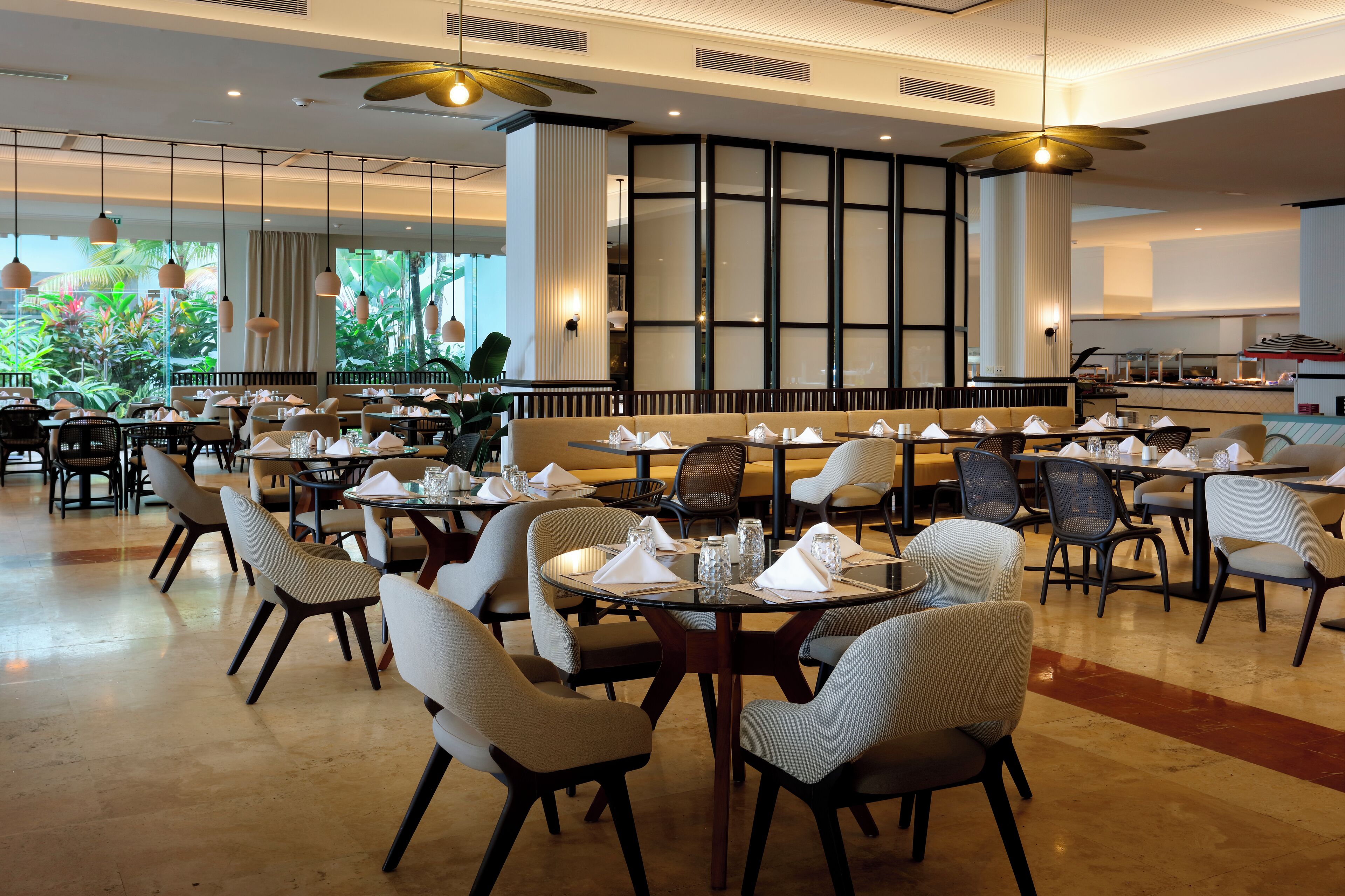 A spacious dining area with multiple tables and chairs, large windows with a view of greenery, and a modern ceiling design.