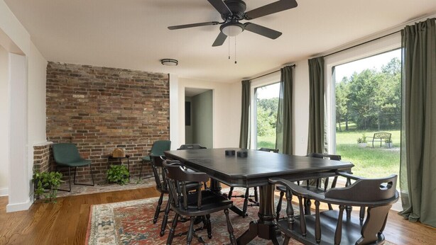 Zona pranzo con parete in mattoni, ventilatore a soffitto, tavolo grande con sedie e vista su un giardino attraverso ampie finestre.