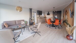 A modern living room with a sofa, coffee table, and chairs, a dining area with orange chairs, and a kitchen in the background. - Runde Panorama - Eden (Heroy)