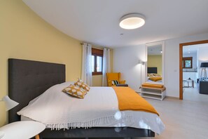 Un dormitorio moderno con una cama grande, una mesita de noche y una silla. Hay una puerta que conduce a otra habitación.