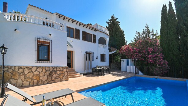 A poolside area with lounge chairs and a table, a stone wall, and a building with a tiled roof and windows. - Apartment Mi Mexico Benissa Costa (Benissa)
