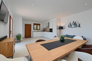 A modern living room with a wooden dining table, white sofas, and a kitchen in the background.