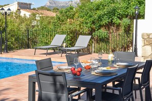 Mesa de comedor dispuesta con platos, vasos y una cesta de frutas, rodeada de sillas, con una piscina y follaje en el fondo.