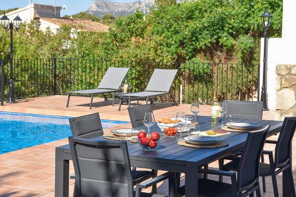 Mesa de comedor dispuesta con platos, vasos y una cesta de frutas, rodeada de sillas, con una piscina y follaje en el fondo.