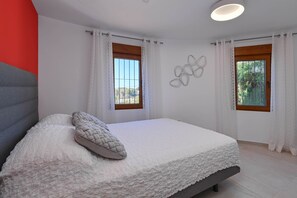 A bedroom with a bed, white curtains, a window, and a wall-mounted light fixture.