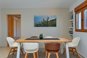 Uma área de refeições com uma mesa de madeira, cadeiras brancas e marrons, um tapete estampado e um quadro na parede retratando uma paisagem.
