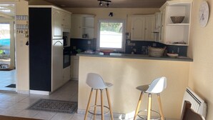 A kitchen with wooden cabinets, a microwave, a coffee maker, and a window providing natural light. There are two white bar stools and a rug on the floor. - Vacation home on enclosed land 800 metres from the beach and shops (Mesquer)