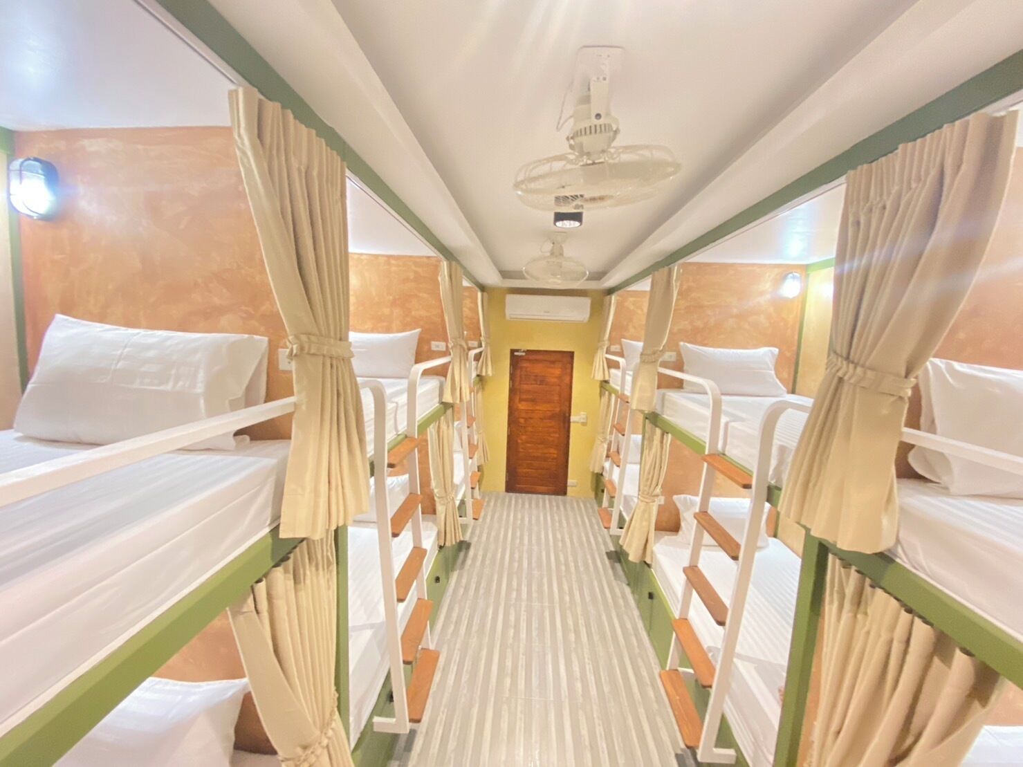 A narrow corridor with bunk beds on either side, a wooden door at the end, and a ceiling fan in the center.