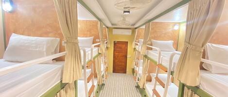 A narrow corridor with bunk beds on either side, a wooden door at the end, and a ceiling fan in the center.