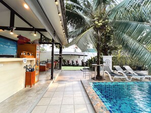 Uma área de piscina externa com espreguiçadeiras, bar e palmeira.
