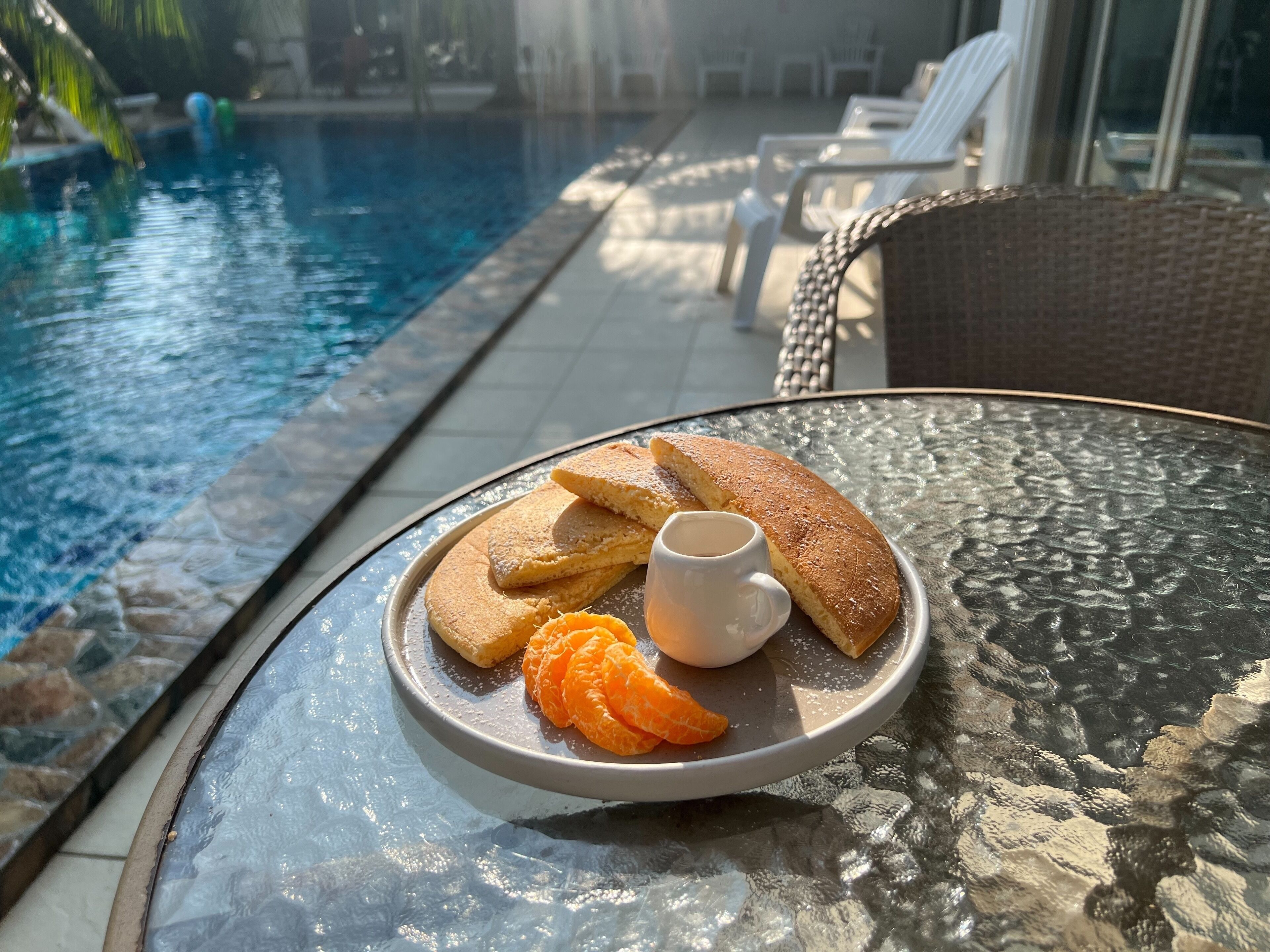 A glass table with a plate of food, a glass of drink, and a pool in the background.