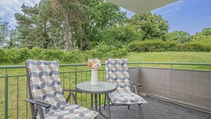 A covered outdoor seating area with two chairs and a table, surrounded by a metal railing and a grassy landscape. - Beautiful vacation apartment in Heringsdorf with terrace - only 400 m to the beach (Seebad Heringsdorf)