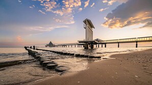 Une jetée s’avançant dans des eaux calmes, avec une structure à son extrémité, une plage de sable et un ciel dégagé.