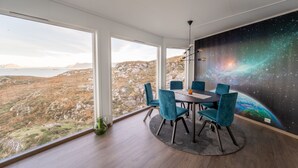 Ein Essbereich mit Tisch und Stühlen, große Fenster mit Blick auf eine gebirgige Landschaft und ein Wandgemälde einer Galaxie.